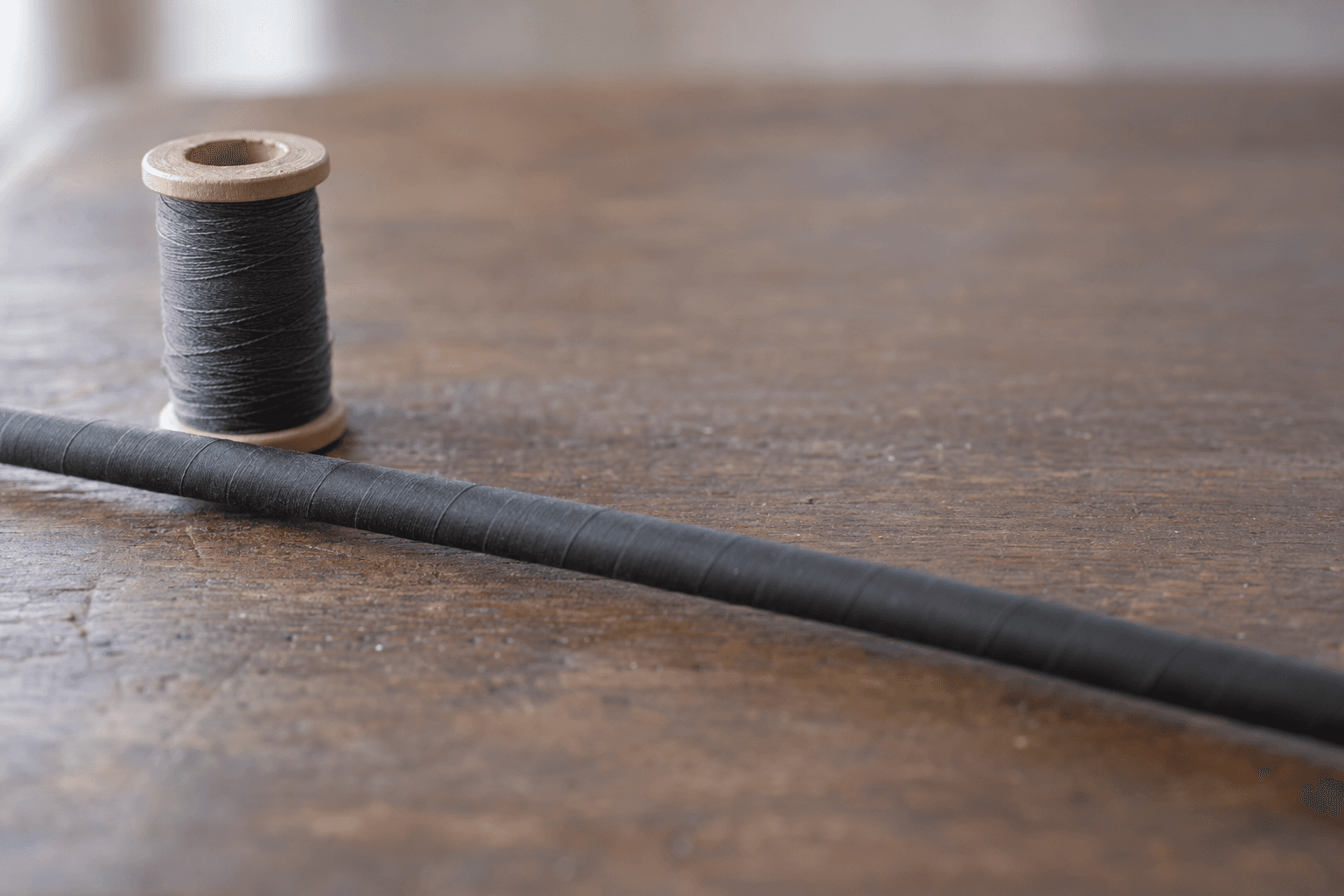 A close-up graphite blank section beside a thread spool on a dusty rod-building bench.
